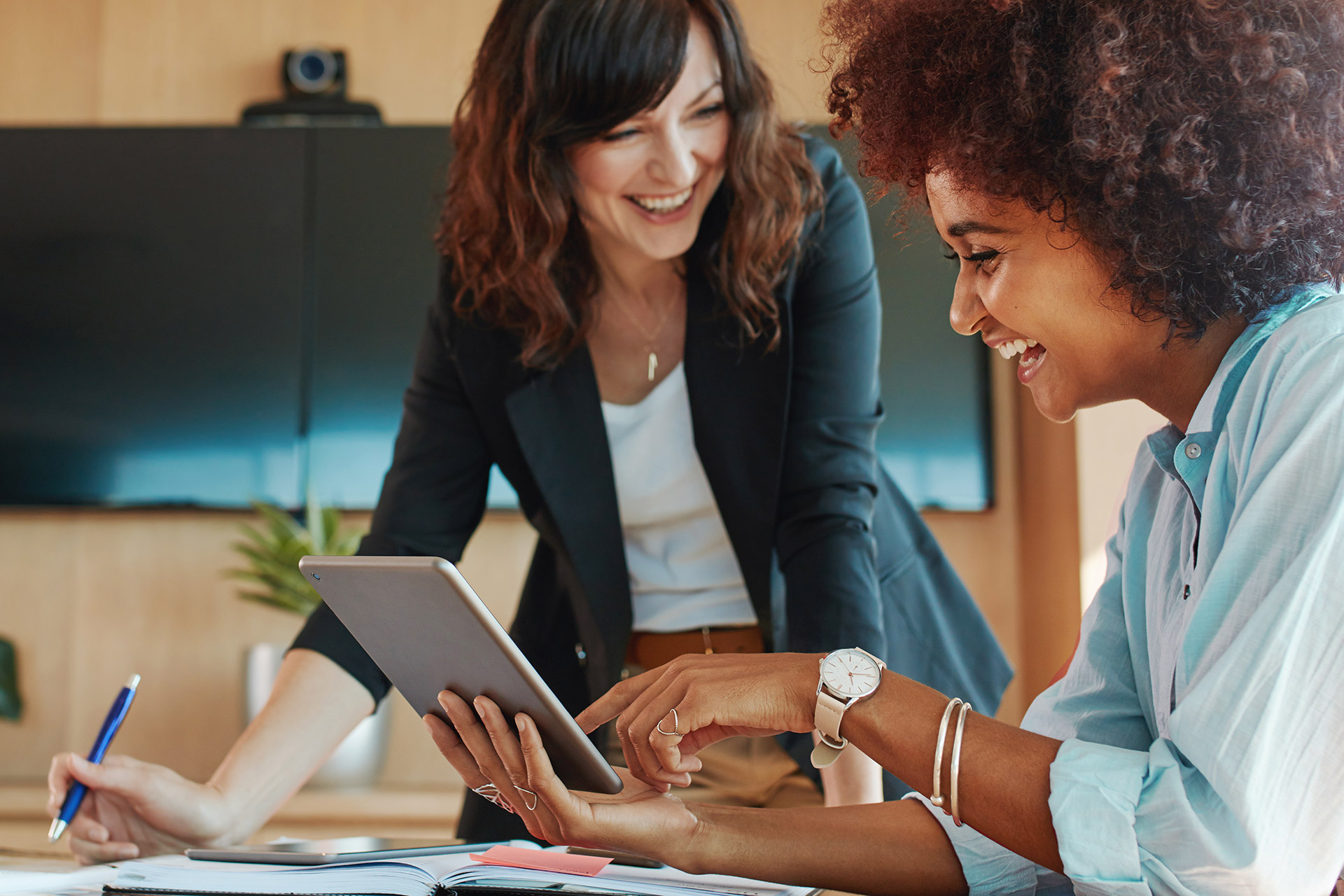 Duas mulheres trabalhando sorridentes em uma mesa. Uma delas com um tablet na mão