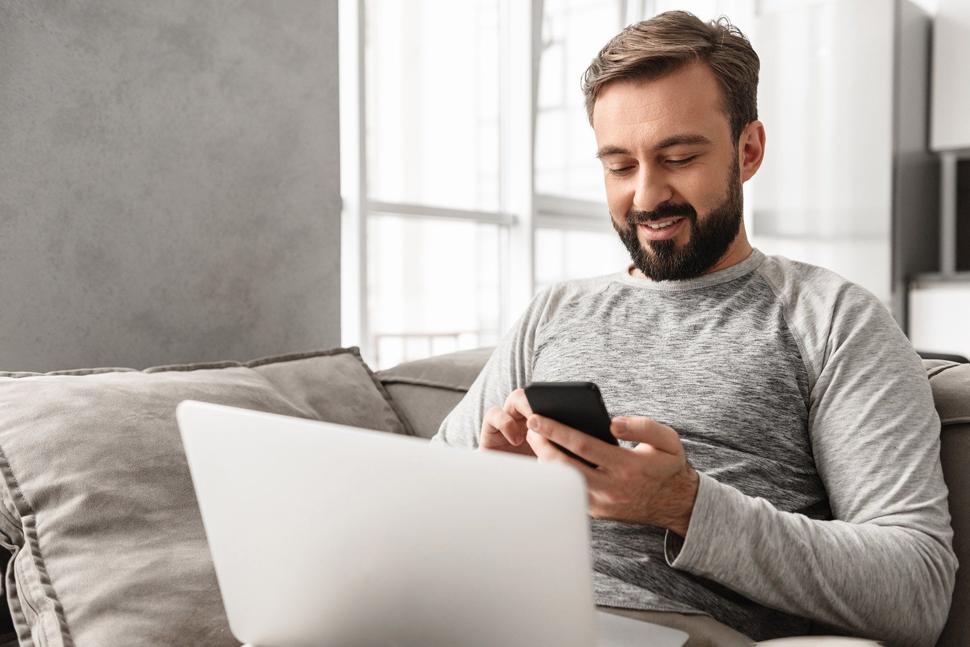 Homem olhando celular na mão com o laptop no colo sentado no sofá