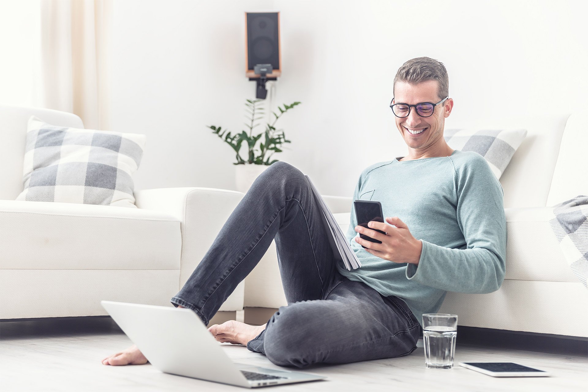 Homem sentado no chão com as costas no sofá usando o celular com o laptop na sua frente, no chão